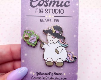 Könnte beinhalten: Ein Emaille-Pin auf einer violetten Cosmic Fig Studio-Trägerkarte. Der Pin zeigt ein weißes Einhorn mit einem schwarzen Hexenhut, das eine grüne, glitzernde Tüte mit der Aufschrift CHIPS und einem kleinen Cupcake-Symbol hält.