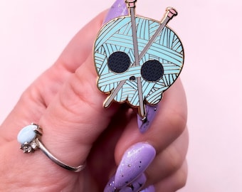 Könnte beinhalten: Ein goldfarbener Emaille-Pin in Form eines Schädels mit einem türkisfarbenen Garnknäuel-Design. Zwei Stricknadeln kreuzen sich über dem Garnknäuel. Der Schädel hat schwarze Augenhöhlen und eine kleine Nase. Der Pin wird von einer Hand mit lila Nägeln gehalten.