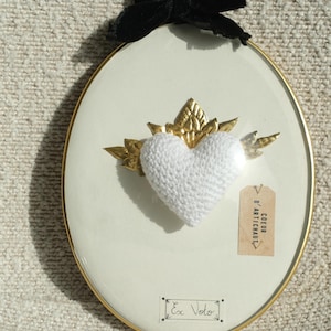 Peut inclure: Un cœur en crochet blanc avec des accents en feuille d'or dans un cadre rond doré. L'image a un ruban de velours noir en haut et une petite étiquette qui dit "COEUR D'ARTICHAUT". L'image a également le texte "Ex Voto" écrit à l'encre noire.