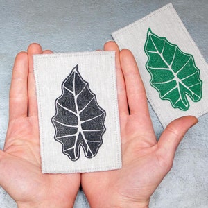 Może przedstawiać: Dwa kwadratowe kawałki tkaniny z wzorem liścia. Jeden kwadrat ma czarny liść, a drugi zielony liść.