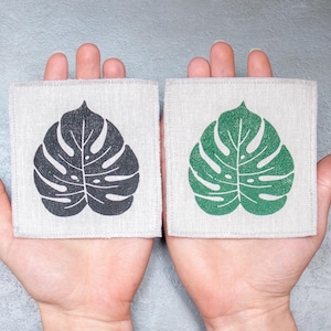 Peut inclure: Deux dessous-de-verre en tissu avec un motif de feuille de monstera. Un dessous-de-verre a une feuille noire et l'autre une feuille verte.