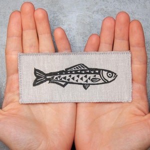 May include: A small, rectangular patch of fabric with a black and white illustration of a fish. The patch is held between two hands.