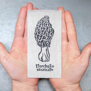 Puede incluir: Un parche de tela blanco con una ilustración en blanco y negro de una seta morchella. El texto "Morchella esculenta" está impreso debajo de la seta.