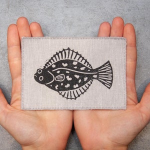 Może przedstawiać: Naszywka z jasnoszarego płótna lnianego z czarno-białym nadrukiem płaskiej ryby.