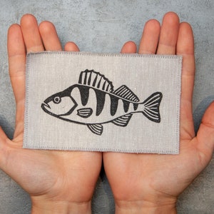 Może przedstawiać: Szary łatkę z tkaniny lnianej z czarno-białym rysunkiem ryby. Łatka jest trzymana między dwoma dłońmi.