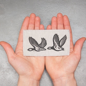 Op de afbeelding: Twee gestileerde vogels in zwart en wit in vlucht, gedrukt op een beige linnen stoffen patch.