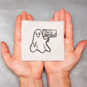 Może przedstawiać: Para rąk trzymająca biały kwadratowy kawałek materiału z czarno-białym rysunkiem ducha trzymającego znak z napisem "This is boo sheet".