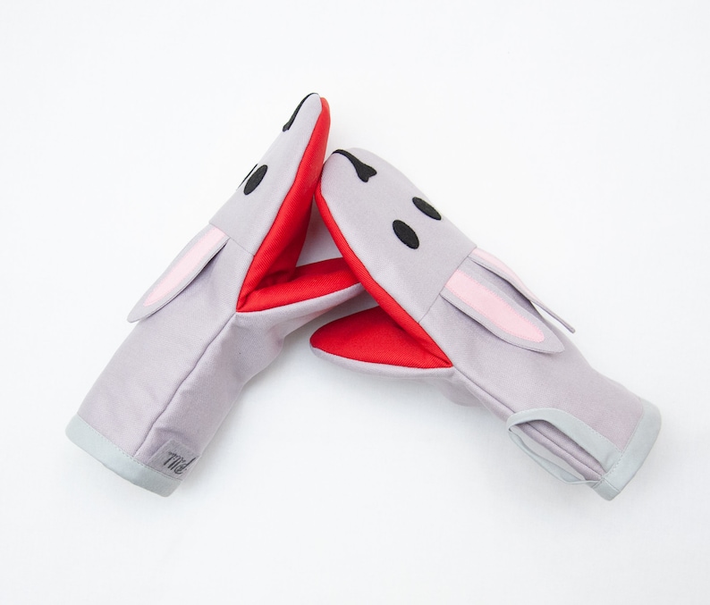 May include: A pair of gray oven mitts with red and pink accents. The mitts are shaped like bunny rabbits with black eyes and a white mouth.