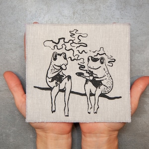 May include: Two black and white frogs sitting on a branch, each holding a cup of coffee. The frogs are facing each other and appear to be enjoying their coffee. The background is a light beige linen fabric.