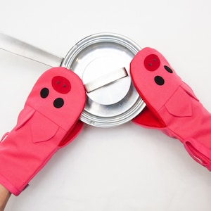 Peut inclure: Une paire de maniques de four roses avec des visages de cochons. Les maniques sont utilisées pour tenir un couvercle de casserole en argent.