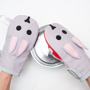 May include: A pair of gray oven mitts shaped like bunnies with pink ears. The mitts are being used to hold a silver pot lid.