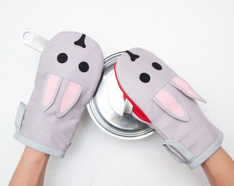 May include: A pair of gray oven mitts shaped like bunnies with pink ears. The mitts are being used to hold a silver pot lid.