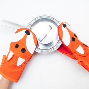 May include: A pair of orange oven mitts with a fox face design. The mitts are being used to hold a silver pot lid.