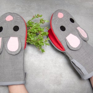 May include: A pair of gray oven mitts with red trim and pink ears shaped like a mouse. The mitts are made of a soft, textured fabric.