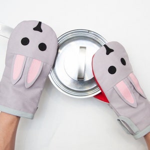 May include: A pair of gray oven mitts with pink bunny ears and black eyes. The mitts are being used to hold a silver pot lid.