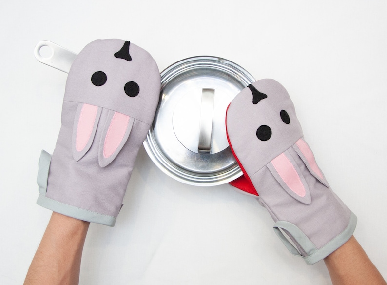 May include: A pair of gray oven mitts with pink bunny ears and black eyes. The mitts are being used to hold a silver pot lid.