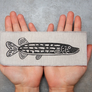 May include: A black and white illustration of a fish printed on a beige linen fabric patch.