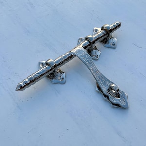 Peut inclure: Un verrou de porte en argent avec un design orné. Le verrou a un corps long et cylindrique avec une extrémité arrondie et une poignée décorative avec un motif floral.