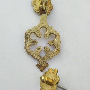 Puede incluir: Un llamador de puerta dorado con un diseño decorativo y ornamentado. El llamador tiene una pieza superior rectangular con bisagras y una pieza inferior en forma de trébol de cuatro hojas. El llamador está sujeto a una puerta con un tornillo.