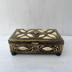 May include: A small, rectangular box with a gold and white design. The box has a hinged lid and sits on four small feet.