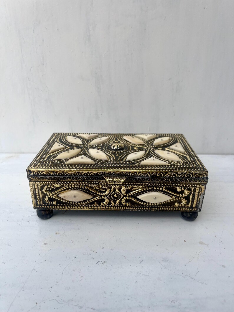 May include: A small, rectangular box with a gold and white design. The box has a hinged lid and sits on four small feet.
