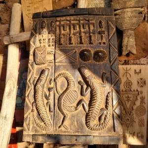 Può includere: Pannello in legno intagliato marrone scuro con intagli a rilievo dettagliati. Il pannello raffigura figure stilizzate, tra cui due coccodrilli che fiancheggiano una forma centrale curva. Ulteriori intagli includono motivi geometrici e figure umane.