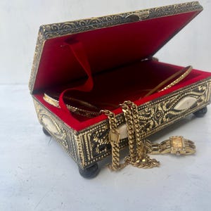 May include: A gold metal jewelry box with an ornate design and a red velvet interior. The box is open and contains a gold chain necklace and a gold watch.