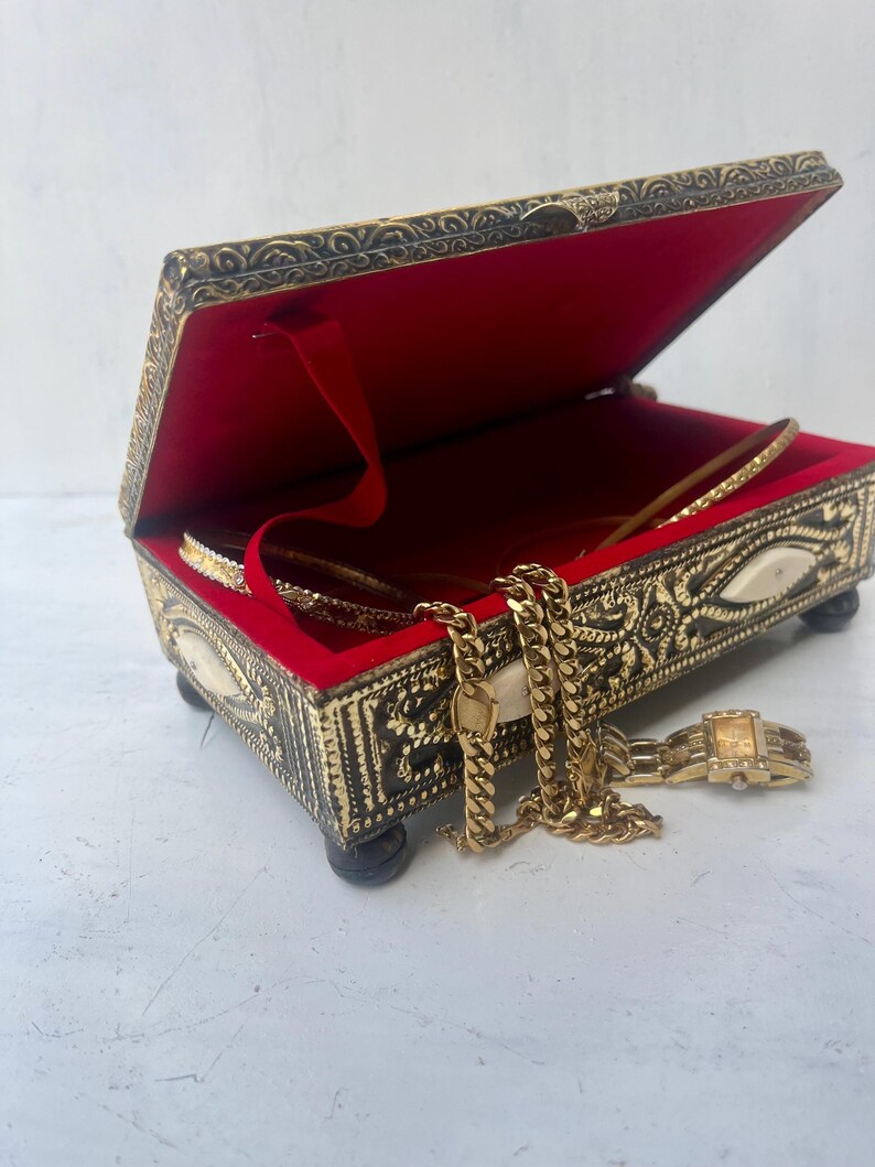 May include: A gold metal jewelry box with an ornate design and a red velvet interior. The box is open and contains a gold chain necklace and a gold watch.