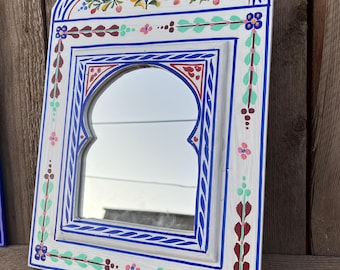 May include: A white painted wooden mirror with a blue and red painted frame. The mirror has an arched top and a floral design painted on the top of the frame.