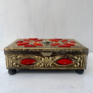 May include: A gold metal box with intricate designs and red accents. The box has a hinged lid and sits on four small feet.