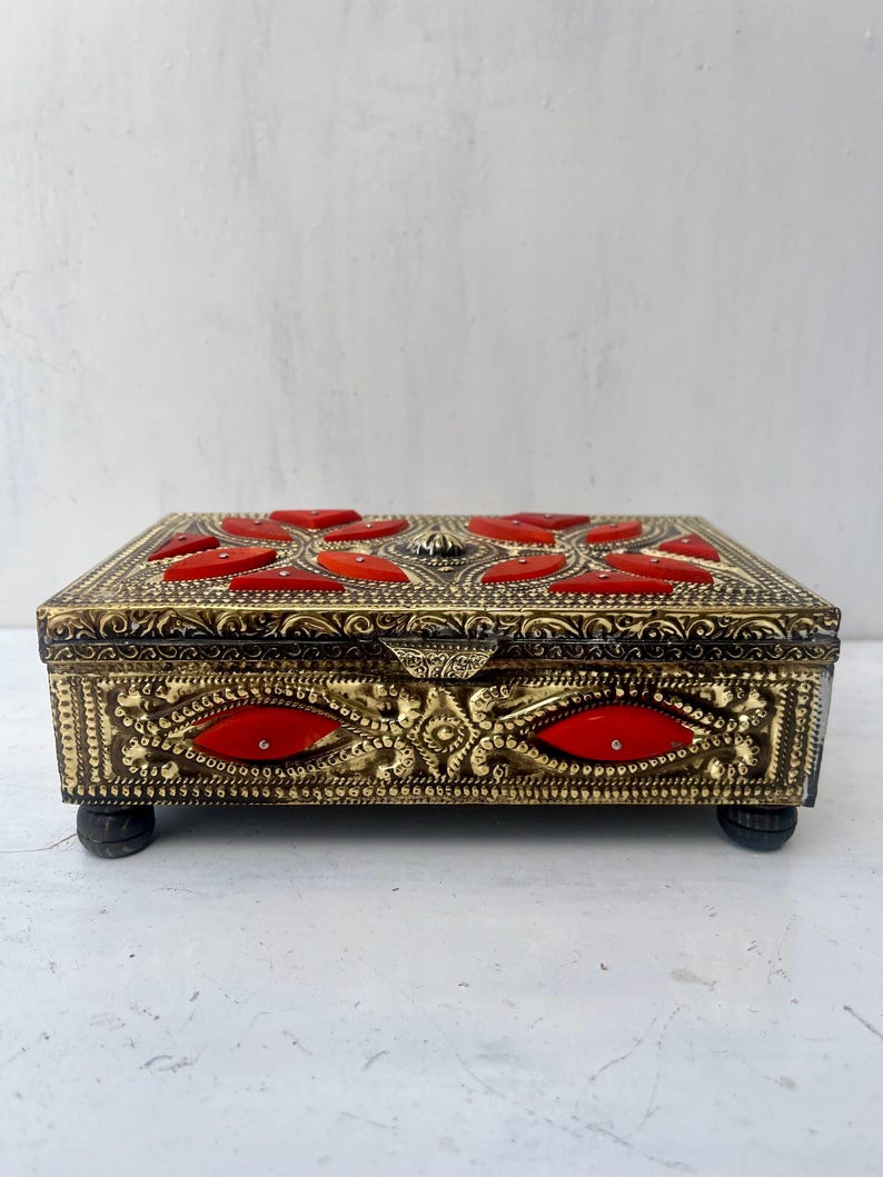 May include: A gold metal box with intricate designs and red accents. The box has a hinged lid and sits on four small feet.