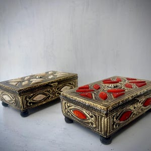 May include: Two ornate decorative boxes with intricate designs. One box is gold with red accents and the other is gold with white accents.