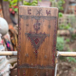 Peut inclure: Une plaque en bois avec des motifs géométriques rouges et noirs complexes. La plaque présente un triangle central avec une lettre stylisée "C" à l'intérieur. Le haut de la plaque comporte trois symboles, dont un "C" et deux autres symboles.