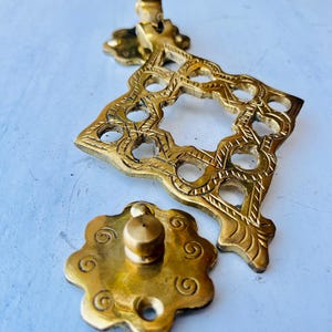 Peut inclure: Une serrure de porte en métal doré avec des motifs complexes et ornés. Le loquet a une forme carrée avec une ouverture circulaire au centre. Le loquet est attaché à une petite pièce décorative ronde avec un bouton en relief.