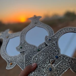May include: Three ornate silver hand mirrors with intricate floral designs and blue and green gemstones. The mirrors are held in a hand against a blurred sunset background.