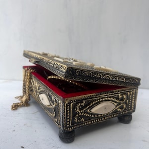 May include: A small, ornate jewelry box with a black and gold finish. The box has intricate carvings and is decorated with white bone inlays. The box is open and has a red velvet interior.