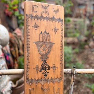 Peut inclure: Une plaque en bois avec un motif sculpté à la main d'une main de Hamsa, symbole de protection et de bonne chance. La plaque est décorée de motifs et de symboles complexes, y compris les lettres "C", "Y" et "U".