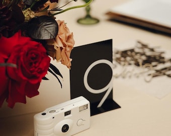 May include: A white disposable camera sits on a table next to a black card with the number "9" written in white. The table is covered in a white tablecloth and there are red and pink flowers in a vase in the background.