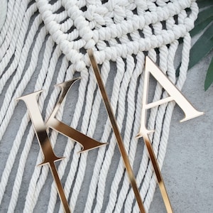 May include: Gold-colored metal letters "K" and "A" on sticks, placed on a white rope backdrop.