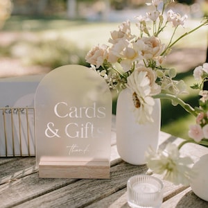 May include: A frosted acrylic sign with a rounded arch top reads "Cards & Gifts" with the word "thank" written below. The sign is on a wooden base and is displayed on a wooden table with a vase of white and cream flowers.