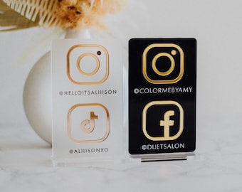 May include: Two acrylic social media signs. One is white with gold Instagram and TikTok logos and handles. The other is black with gold Instagram and Facebook logos and handles. The signs are displayed on a white surface.