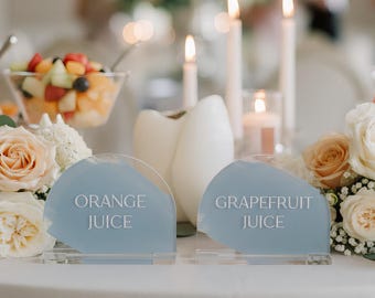 Può includere: Due insegne semicircolari in acrilico azzurro con testo bianco che recita ORANGE JUICE e GRAPEFRUIT JUICE, posizionate su un tavolo con composizioni floreali bianche, candele e ciotole di vetro riempite di macedonia di frutta fresca.