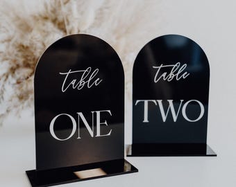 May include: Two black acrylic table number signs with rounded tops. The signs have white text that reads "table one" and "table two".
