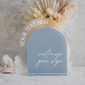 May include: A frosted acrylic sign with a semi-circle shape. The top half is a light brown color with the text "YOUR TEXT HERE" in white. The bottom half is a light blue color with the text "customize your sign" in white.