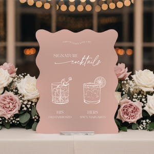 May include: A pink, wavy-edged sign with the words "Signature Cocktails" in elegant script. Below, illustrations depict "His" Old Fashioned and "Hers" Spicy Margarita cocktails. The sign is set on a white tablecloth, surrounded by floral arrangements.