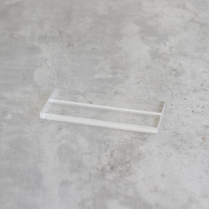 May include: A clear acrylic display stand with two shelves. The stand is designed to hold small items, such as jewelry or other collectibles.