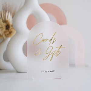 May include: A frosted acrylic sign with a gold inscription that reads "Cards & Gifts" and the text "Thank You!" in black below. The sign is on a white surface.