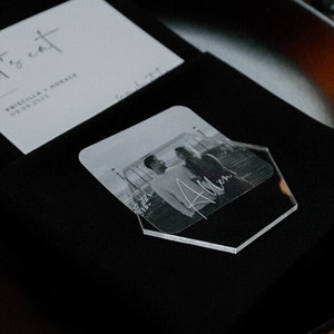 May include: A black and white photo of a couple is displayed on a clear acrylic hexagonal place card with the name "Allen" printed on it. The place card is on a black napkin on a black plate. The text "Let's eat" is on a white card next to the place card.