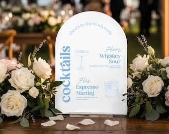 May include: A white arched sign with the word "cocktails" in light blue, listing "Hers Whiskey Sour" and "His Espresso Martini" drinks. The sign is on a wooden table, surrounded by white and pink roses, with a blurred wedding scene in the background.