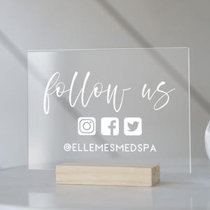 May include: A clear acrylic sign with the text "follow us" in white cursive font. Below the text are icons for Instagram, Facebook, and Twitter. The sign also includes the text "@ELLEMESMEDSPA" in white.